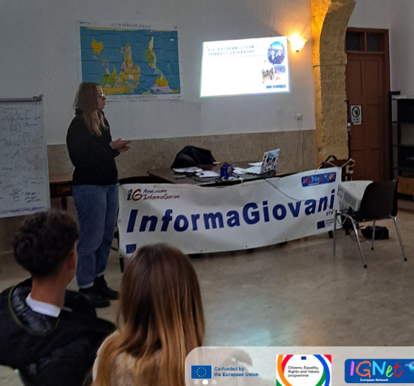 Una volontaria di InformaGiovani presenta le opportunità europee a un gruppo di giovani, con slide proiettate e lo striscione dell'associazione in primo piano