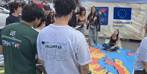 Volontari di InformaGiovani e giovani del quartiere partecipano a un'attività all'aperto attorno a un grande telo con la mappa dell'Europa, davanti allo stand di Europe Direct Palermo, durante le celebrazioni della Festa dell'Europa.