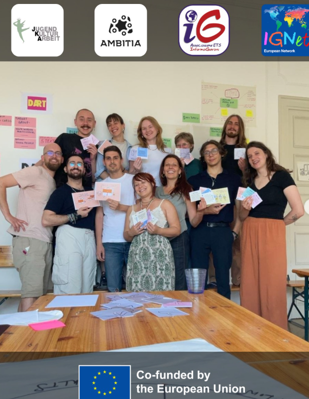 I partecipanti al meeting di coordinamento del progetto DART – Digital Awareness Reflection Toolkit posano sorridenti mostrando le carte dello strumento in sviluppo, davanti a una parete piena di post-it e cartelloni di lavoro