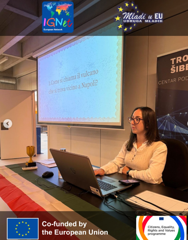 Una conduttrice guida il quiz interculturale sulla cultura italiana dalla postazione decorata con il tricolore, con la prima domanda proiettata sullo schermo (