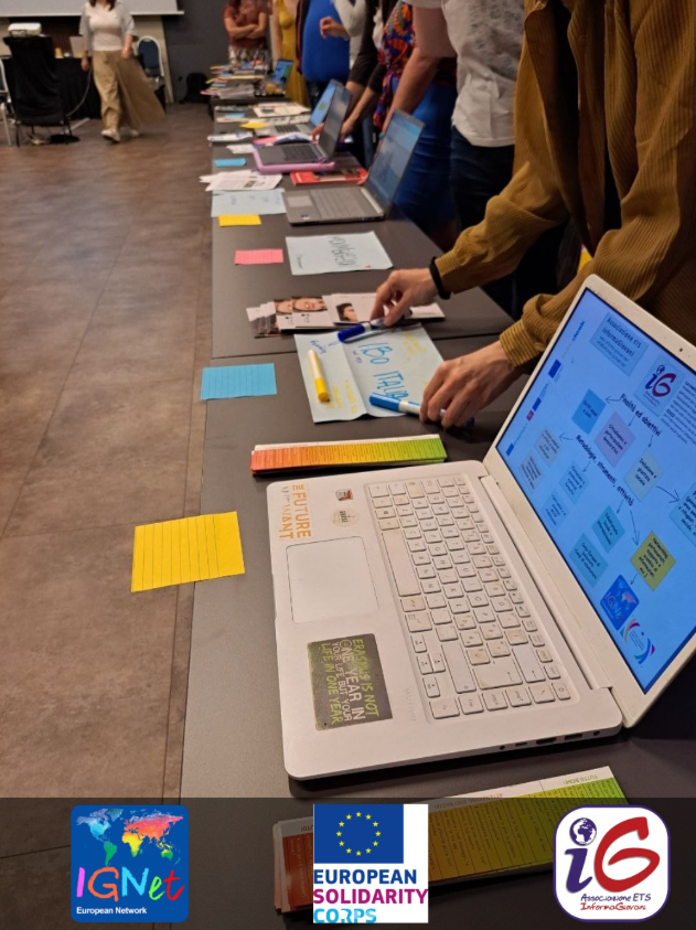 Il tavolo espositivo del training nazionale per le organizzazioni del Corpo Europeo di Solidarietà a Roma, con computer portatili, brochure e post-it colorati delle realtà partecipanti; in primo piano lo schermo con la mappa delle attività di InformaGiovani ETS e un adesivo con la scritta 