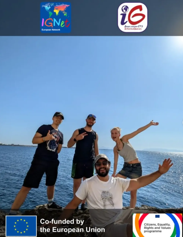I quattro volontari internazionali del progetto di Castelvetrano posano sorridenti e in pose scherzose sugli scogli, con il mare della costa siciliana e il cielo limpido alle spalle, durante un momento di tempo libero; in alto i loghi della rete IGNet e di InformaGiovani ETS, in basso quelli del cofinanziamento dell'Unione Europea e del programma CERV.
