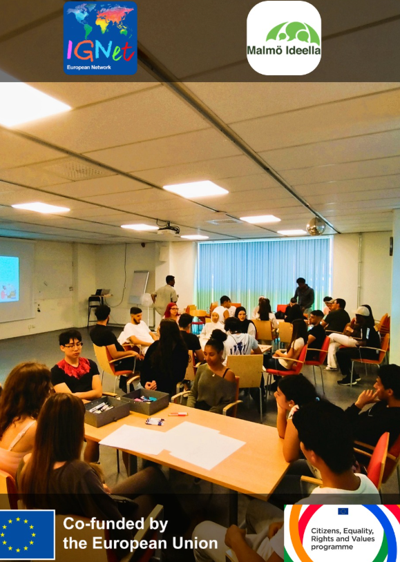 Un gruppo numeroso di giovani siede in cerchi di sedie e attorno a un tavolo con fogli bianchi e pennarelli, in una sala con schermo di proiezione e lavagna a fogli mobili.
