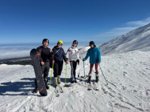 quattro persone su una montagna innevata, in una giornata limpida con cielo azzurro. Tre di loro indossano sci e tengono i bastoncini, mentre la quarta persona tiene una tavola da snowboard in posizione verticale. Tutti indossano abbigliamento da sport invernali, come giacche, pantaloni tecnici e caschi. Sul fondo si vede un ampio paesaggio di neve, con creste montuose e nuvole basse all’orizzonte. L’atmosfera è quella di un’attività sportiva all’aperto in un ambiente alpino.