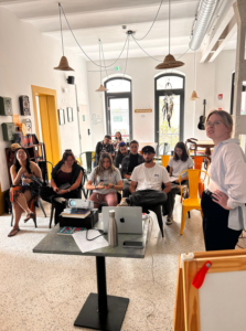 L’immagine mostra un gruppo di persone sedute in una piccola aula durante un incontro formativo. I partecipanti sono rivolti verso un tavolo su cui si trovano un computer portatile, una borraccia, alcuni fogli e un telefono. Una persona in piedi conduce la sessione. La stanza ha pareti chiare, grandi finestre, lampade con paralumi intrecciati e scaffali con libri e oggetti decorativi. 