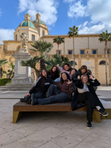 sette ragazze sedute in posa su una panchina con alle spalle un palazzo storico