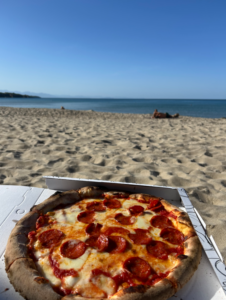 Una pizza in scatola è appoggiata sulla sabbia, con il mare e il cielo limpido sullo sfondo. Alcune persone sono distese vicino all’acqua.