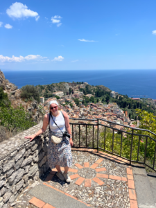 Una persona è in piedi su una terrazza che affaccia su una città costiera costruita sul pendio. Si vedono edifici con tetti arancioni, alberi e il mare blu sotto un cielo sereno.